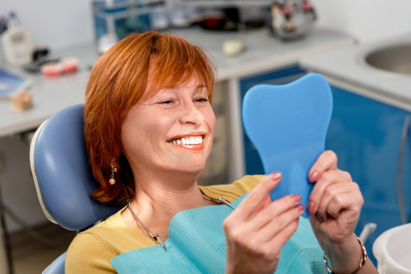 Older woman smiling with teeth into a handheld mirror while sitting in a dental chair